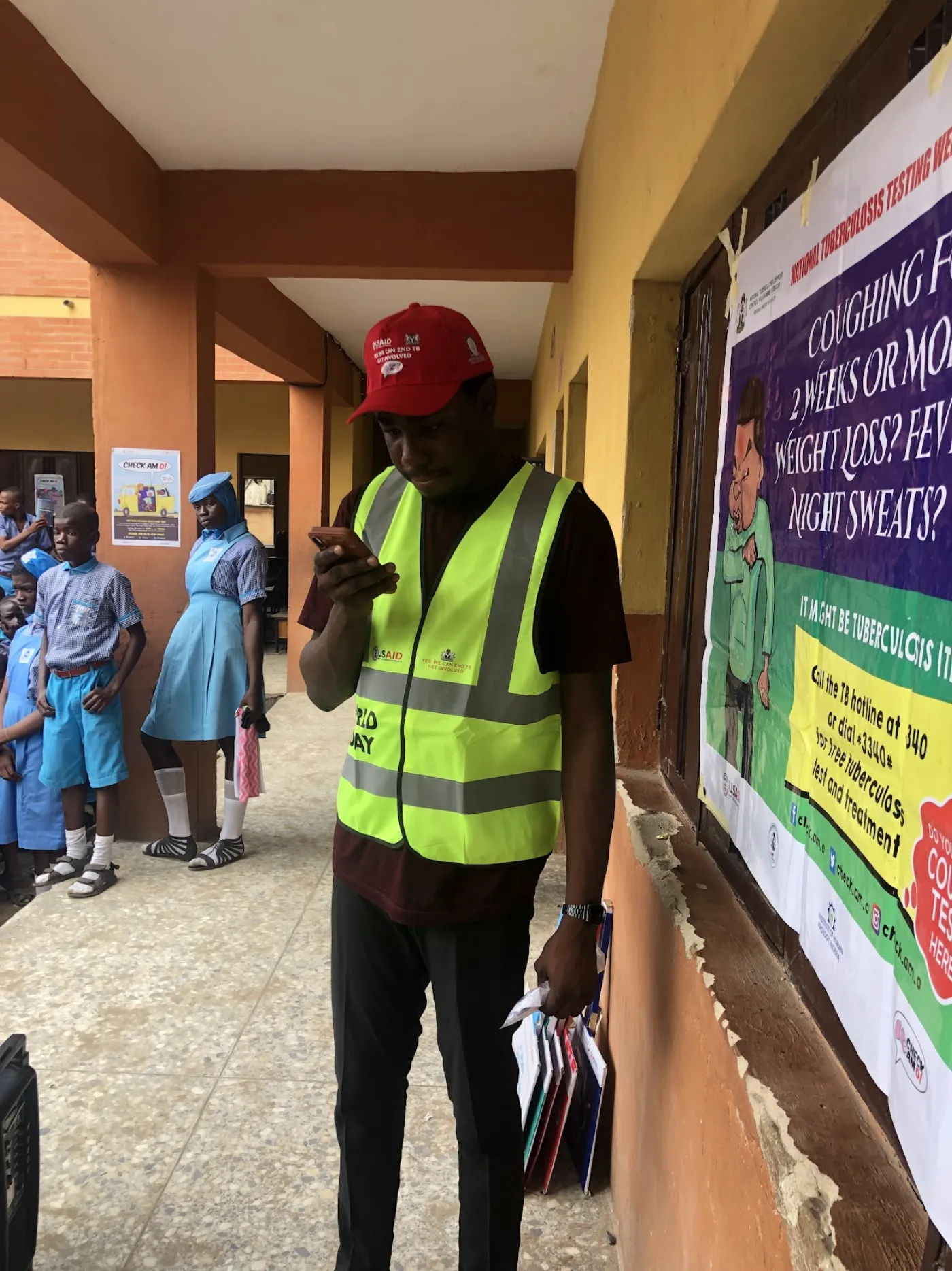 Tomiwa in a World TB Day volunteer vest beside a tuberculosis symptom-awareness poster at a Lagos school